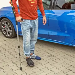 Mężczyzna w ortezie na nodze opiera się o laskę, gotowy do jazdy samochodem.