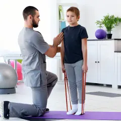 Fizjoterapeuta pomaga chłopcu w ćwiczeniach z gumą. Rehabilitacja wad postawy u dzieci NFZ to szansa na zdrowszy kręgosłup.