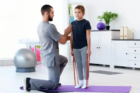 Fizjoterapeuta pomaga chłopcu w ćwiczeniach z gumą. Rehabilitacja wad postawy u dzieci NFZ to szansa na zdrowszy kręgosłup.