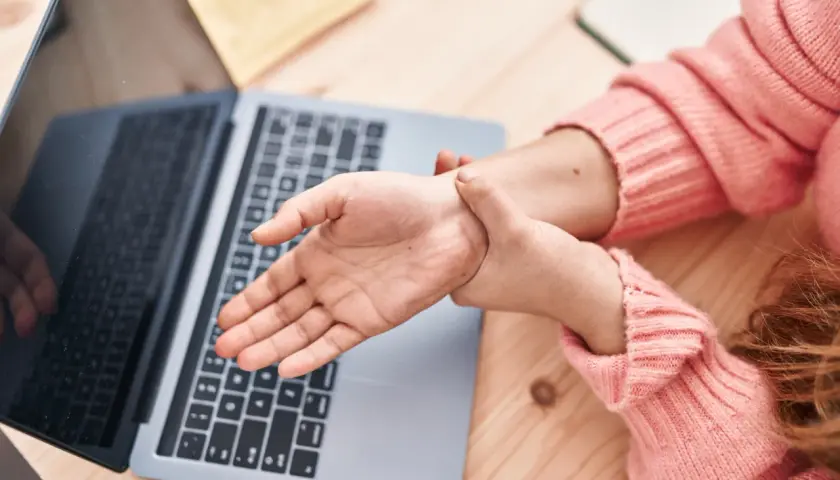 Dłoń masuje nadgarstek przy laptopie, szukając domowych sposobów leczenia cieśni nadgarstka.