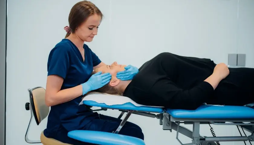 Fizjoterapeuta szczękowy delikatnie masuje twarz pacjentki leżącej na kozetce.