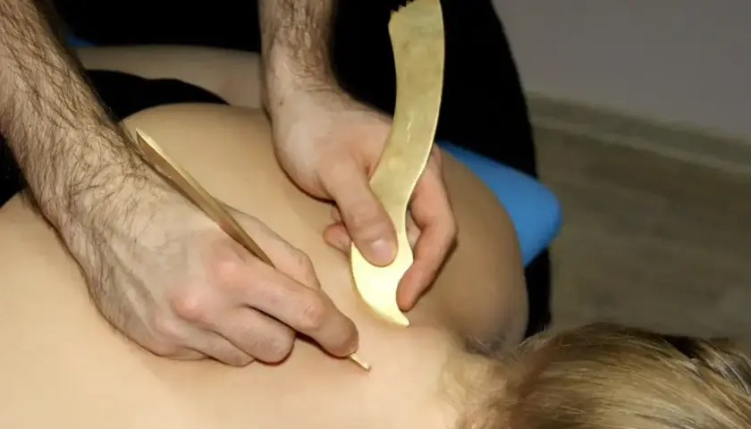 Masaż narzędziami do pinoterapii. Terapeuta pracuje nad karkiem pacjenta, używając złotych przyrządów do głębokiego masażu tkanek.