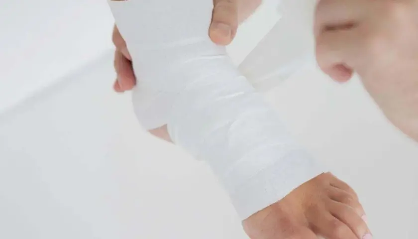 Dłonie owijają stopę białą taśmą do tapingu, zapewniając wsparcie i stabilizację.