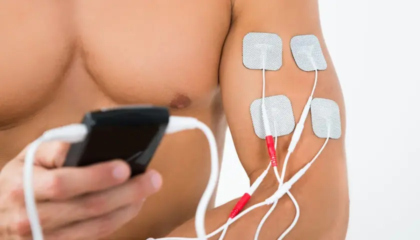 Mężczyzna stosuje elektrostymulację TENS na ramieniu. Elektrody podłączone do urządzenia to element fizjoterapii.