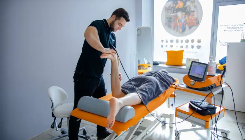 Fizjoterapeuta wykonuje zabieg falą uderzeniową na stopę pacjenta.