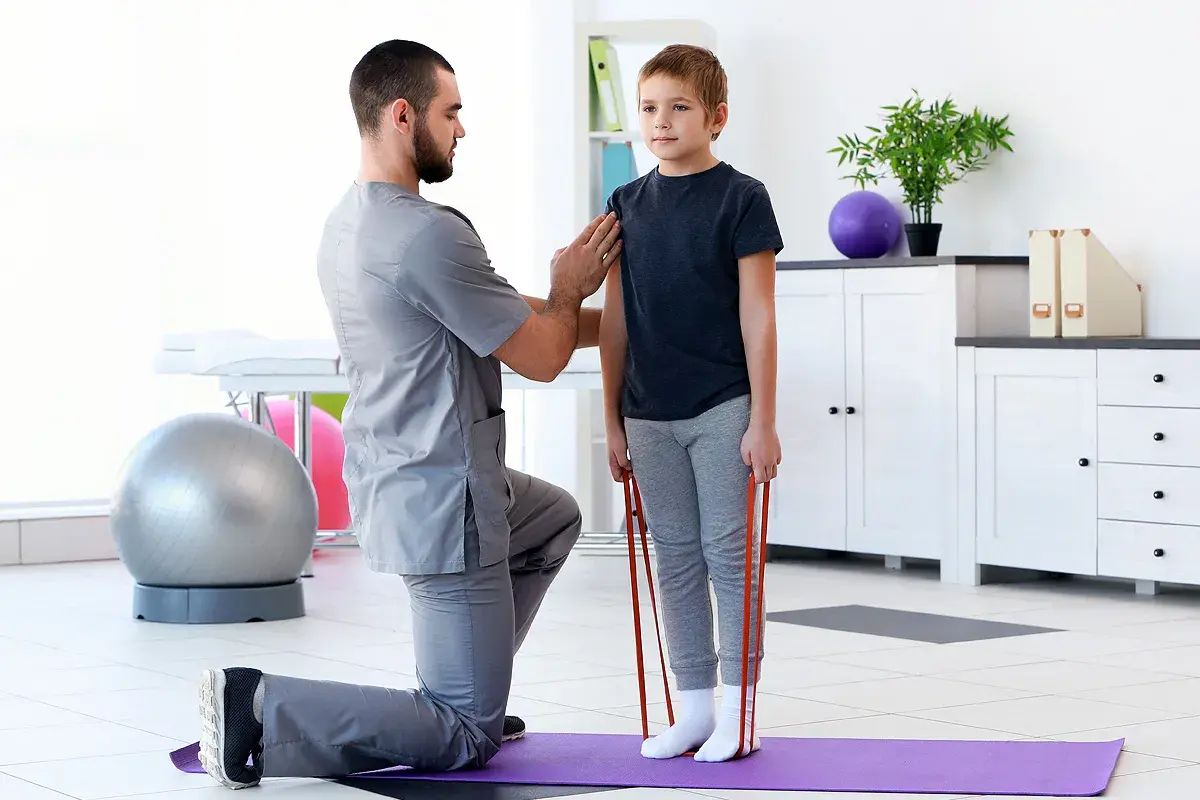 Fizjoterapeuta pomaga chłopcu w ćwiczeniach z gumą. Rehabilitacja wad postawy u dzieci NFZ to szansa na zdrowszy kręgosłup.