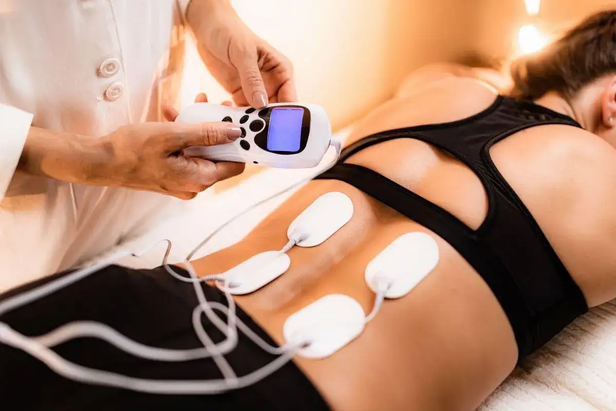 Fizjoterapeuta ustawia aparat do elektrostymulacji TENS na plecach pacjentki. Elektrody podłączone do urządzenia mają przynieść ulgę w bólu.
